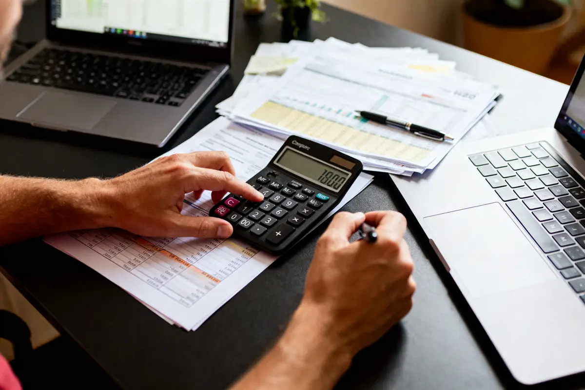 Personne utilisant une calculatrice et un stylo, documents financiers, deux ordinateurs portables sur un bureau.